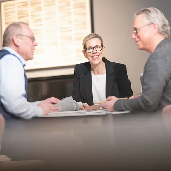 Drei Personen sitzen am Tisch und betrachten Stoffmuster, Beraterin in der Mitte, zwei Kunden gegenüber